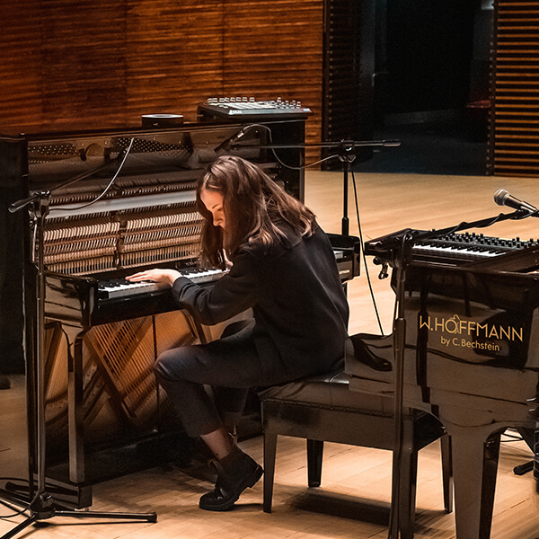 Pianistin Hania Rani sitzt am schwarzen geöffneten Klavier, hinter sich ein schwarzer Flügel in Konzertsaal